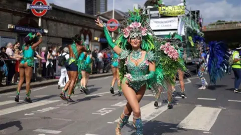 PA a woman dressed in a green and blue peacock-inspired carnival costume, dances in front of the camera with other dancers in turquoise costumes behind her, next to Westbourne Park station. A carnival float is visible but blurred in the distance
