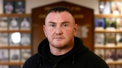 Ross is wearing a dark hoody and standing in the middle of a rugby club, facing the camera.