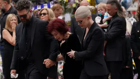 PA Sharon Osbourne is supported by her son and daughter has they see the flowers and memorabilia left for Ozzy by his bench.
