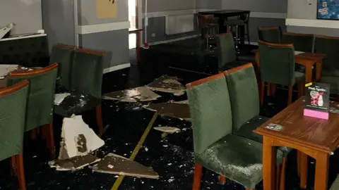 Pauls Place Broken ceiling panels on the floor of a community centre in Yate, with green chairs and tables covered in dust.