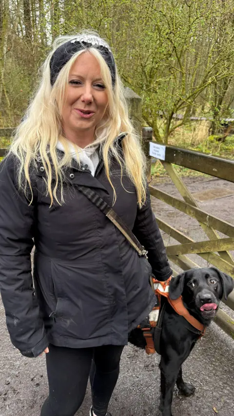Woman in a dark coat standing by a wooden gate in woodland, with a black dog in an orange harness looking up.
