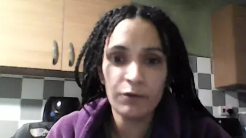 Ayshea Malik, with her black hair in braids and wearing a purple jumper, speaks to the camera sitting in a kitchen with cupboards and a work surface behind her