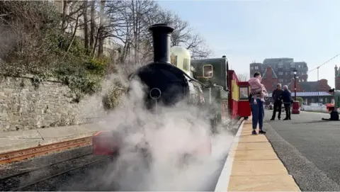 Front of a steam train at Douglas station, people standing on the platform, steam coming from the front car