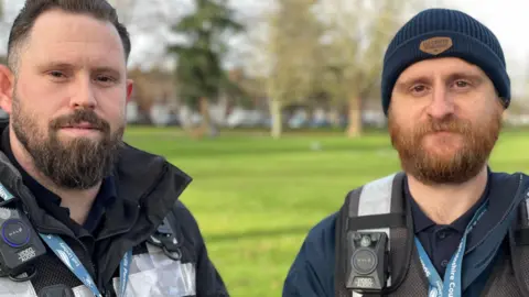 A man with a black beard and another man with a ginger beard both wearing high vis vests and body cameras smile at the camera