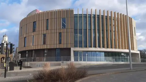 LDRS The outside of The Word which is a large, round, modern building with wooden panels lining the windows on the upper floors. There is a large glass entrance on the bottom floor and a glass railing in front of it.