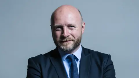 Liam Byrne Liam Byrne wearing a navy suit and, light blue shirt and blue tie. He is standing against a grey backdrop
