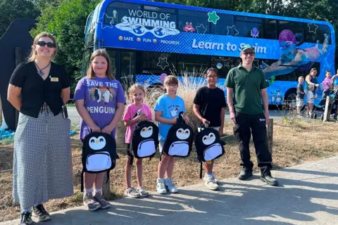 Children and staff pose with penguin backpacks in front of the World of Swimming bus.