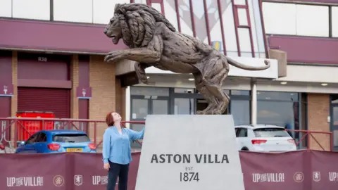 A large bronze lion statue that is standing on its back legs and looks like it is about to pounce forwards. It is on top of a grey platform that reads "Aston Villa, EST 1874". A woman in a blue shirt is standing next to the platform and looking up to the lion. She has red, short hair. The statue is in front of a football stadium and two parked cars are behind a barrier. 