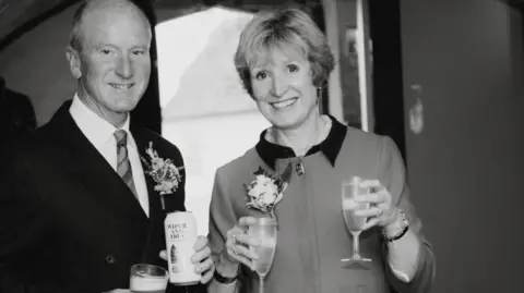 Photo of Tacey and David at a wedding. It is a black and white photo. David is wearing a suit and tie and holding a can or beer and a glass of beer in both hands. Tacey is smiling to the camera holding wine glasses in both hands. 