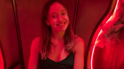 Handout Libby smiling into the camera with her teeth. She looks to be in a bar with red lights around her. Her hair is down and she is wearing a black top and the same simple necklace.