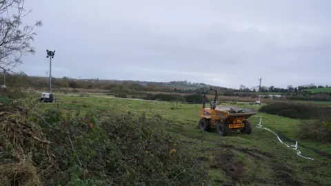 PA Media The image shows a countryside landscape of green fields and bushes in the foreground. Off to the right of the image is a yellow digger-type piece of machinery, to the left is a grey coloured flood light that is turned off. The sky is a slate grey colour. 