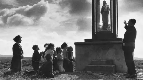 Father John Morrison talks to some of his parishioners next to a shrine. The parishioners are made up of a group of children, who are accompanied by a woman. The group are on their knees.