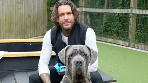A man with shoulder-length brown hair sitting with a grey dog. The dog is a Neapolitan mastif. They are in a garden at a Dogs Trust home. The man has tattoos on his arms and is wearing a white top and black gilet. 