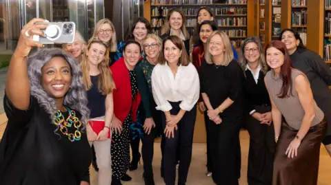 DSIT Anne-Marie Imafidon takes a selfie on her smartphone of a crowd of women behind her. Technology secretary Liz Kendall stands in the centre, wearing a white shirt and black trousers, and smiling.