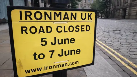 A yellow road sign reading: 'IRONMAN UK, ROAD CLOSED 5 JUNE TO 7 JUNE'.