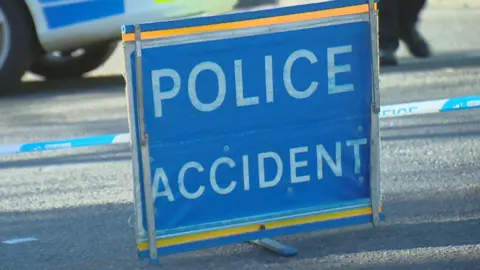 BBC A blue 'POLICE ACCIDENT' sign on a road, with police tape behind it, and a police vehicle.