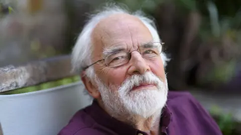 John Marjoram staring into the camera smiling. He is wearing a purple shirt and glasses. He is sitting on a bench in a garden setting.