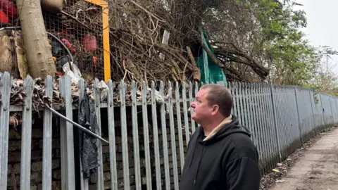 Simon Thake/BBC A man in a black hoodie looks up at a silver metal fence which appears to be holding back piles of rubbish.