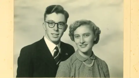 Family handout Mary, on the right, wearing checked blouse with what looks like a double string pearl necklace. Her hair is short and styled. She is with her husband Philip, on the left, wearing round glasses and a suit jacket, shirt and tie. They are both looking at the camera, in a posed photograph. The image is in black and white.