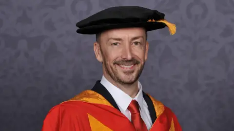 A man wears a university gown which is orange and yellow over his dark suit. He has a beard and and is also wearing a black cap with a yellow tassel.