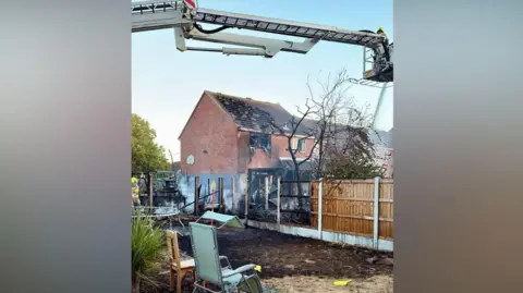 Essex County Fire and Rescue Service Part of a house roof is burnt, along with a window. Burnt garden fences and fire debris as well as smoke are evident. A fire service hydraulic ladder is above one of the gardens.