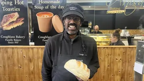 ADRIAN HARMS/BBC Romesh Ranganathan wearing a black cap and black overall and holding a loaf of bread standing behind the counter at Coughlans Bakery in Dorking