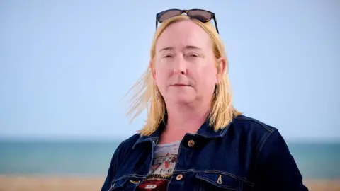 Jim Holden / BBC A woman with blonde shoulder -length hair that stands on a pebble beach with the sea in the background. She wears a blue denim jacket. The sky is blue and cloudless.