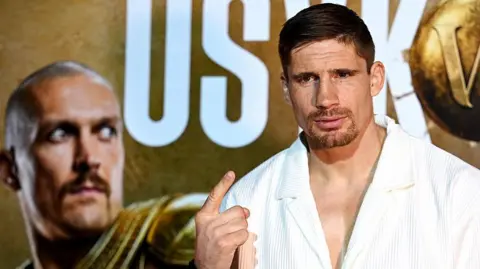 Rico Verhoeven holds up one finger and stands in front of a board with an image of Oleksandr Usyk
