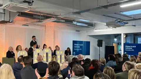 Martin Birch, director of children and family wellbeing at Cumberland Council, standing on a stage with other members of the children's services team, in front of a crowd of the department's staff. A group of children holds letters that light up, spelling the word 'Good'.