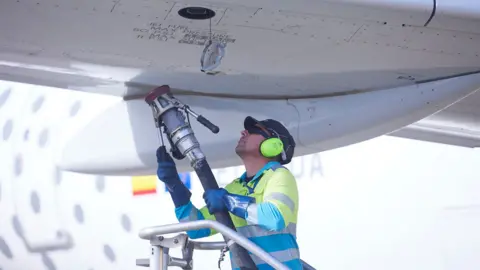 A person wearing hi vis and ear defenders lifts a jet refueling pump towards the fuel port of the wing of an aircraft on a sunny day at Seville airport in 2022.