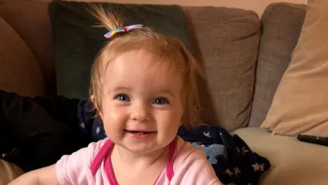Amy Sutton A baby girl is smiling while sitting on or near to a cream coloured sofa. Her lower two front teeth can be seen. She is wearing a pink baby grow and her short blonde hair is tied in a tiny ponytail with a rainbow clip.