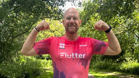 A bald man wearing a pink sports shirt grins at the camera as he flexes his arms. The shirt has the Cure Leukaemia logo on it.