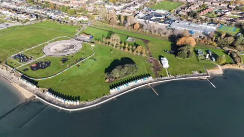 A drone photo of a large grassy park next to the sea. There's a road and a row of beach huts separating the park from the sea.