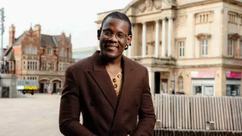 A man wearing a brown coat and knitted top stands in a town square.