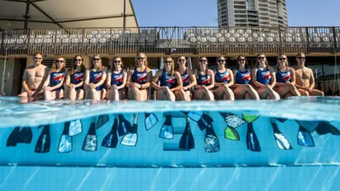 CMAS A group of people in red, white and blue swimming suits sat by the edge of an outdoor pool. The group are sat wearing flippers in the water. Half of the shot is above the water, while the other half is underwater.