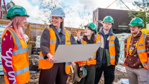 Merlin Grian, Aimsey, DanTDM and LDShadowLady wear orange hi-vis vests and green safety helmets with Minecraft World logos on them. A woman also wearing safety gear shows them a large card with plans for the park. They are surrounded by building equipment and materials.