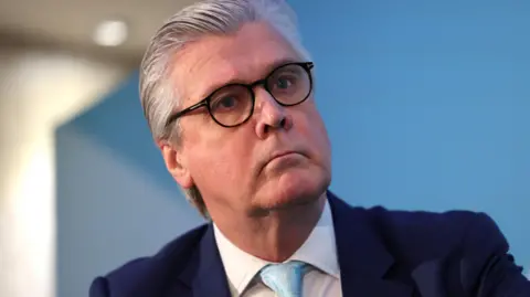 Malcolm Offord, who has grey hair brushed back, in a close-up shot in front of a pale blue background.
