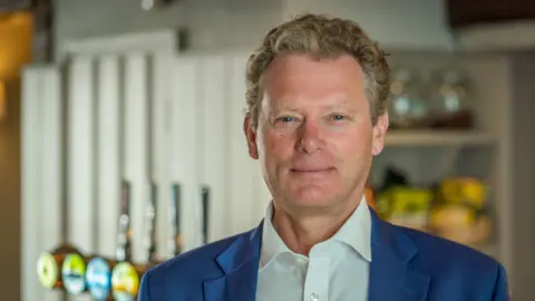 PA Media Jonathan Neame, smiling and looking into the camera while wearing a suit and standing in a bar