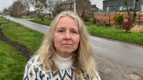 John Devine/BBC Denise Pearce has shoulder length blonde hair, parted in the middle. She is wearing a white woollen jumper with blue and black diamond patterns on it.
Behind her is a single track road with some buildings visible and a grass verge on the left and right with some trees growing.