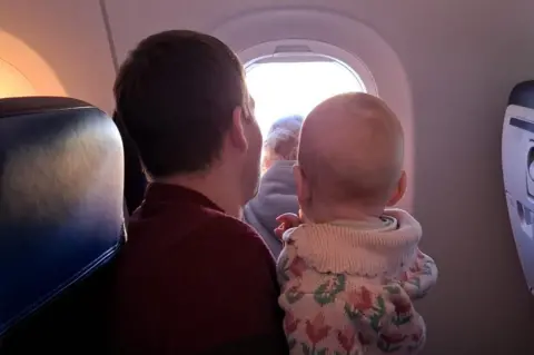 Sarah Rodgers A man sits on an aeroplane holding a baby on his lap as they both look out of the window at the light outside.