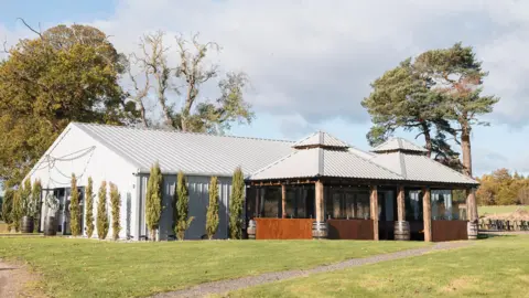A large barn made of steel and wood sits in a large green expanse, surrounded by trees 