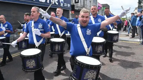 Pacemaker Archive photo: Band members parading through Ballymena on 12 July 2024. The men are wearing blue polo shirts with a band crest on their chest, black trousers and white gloves and they are each carrying navy blue drums via a strap over one shoulder. One drummer, Stephen Steele, is holding his drumsticks in outstretched arms and is smiling or gesturing at the camera.