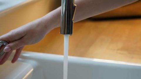 A woman's hand turning on a chrome tap of a belfast-type kicthen sink with part of a wood-effect worktop in the background.