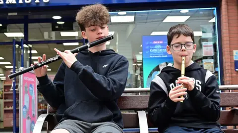 Two young boys with brown hair, wearing black hoodies, are playing wind instruments on a bench 