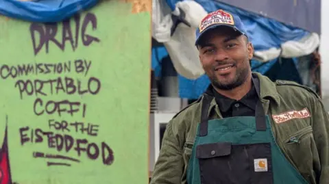 BBC Cassius Walker Hunt wears a khaki green jacket with San Portablo, the name of his business, embroidered on it and a green and black Carharrt apron and blue cap. He is pictured in front of his coffee van which has a red dragon painted on the side and the words Draig, commission by Portablo Coffi for the Eisteddfod