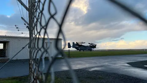 A military aircraft on a runway, photographed through a chain-link fence. 
