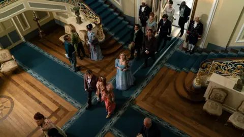 Courtesy of Netflix Interior scene of a grand staircase hall with patterned blue carpets, polished wood floors, and ornate railings. People in period dress are walking inside, roughly in a line. The photo is taken from above.
