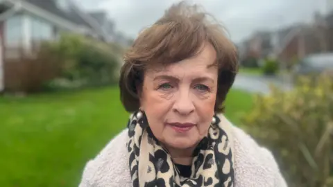 A woman with short brown hair standing outside in front of a garden. She is wearing a scarf and coat.