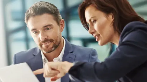 Getty Images A man in a white shirt and blue jacket looks at a piece of paper while a woman in dark jacket points at the paper.