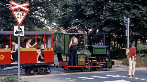 Chris Grimes A woman standing in a road, holding a red flag, wearing a hat, with a train, with passengers in it, crossing a road. There are two road signs and trees behind the train.